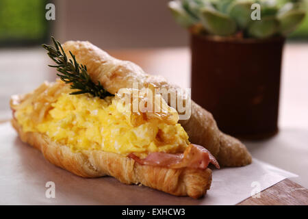 Auf eine Holzplatte auf einem Holztisch serviert leckere Croissant mit Speck, Rührei Ei karamelisiert Zwiebeln und Rosmarin. Stockfoto