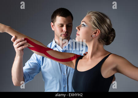 Schöne weibliche Ballett-Tänzerin und ihr Physiotherapeut auf grauem Hintergrund. Ballerina trägt einen schwarzen Anzug und hat rote st Stockfoto