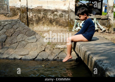 wenig junge sitzen allein auf kleine Steinbrücke über Wasserweg mit einem Stück Schnur Fischen in einem Dorf in der Nähe von Solo Java spielen Stockfoto