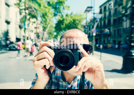 ein junger kaukasischer Mann mit Sonnenbrille zeigt seine Kamera auf den Betrachter auf La Rambla in Barcelona, Spanien Stockfoto