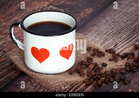 Eine Tasse schwarzen Kaffee in eine alte Emaille Becher mit Herzen, Kaffeebohnen auf Vintage rustikales-Hintergrund Stockfoto