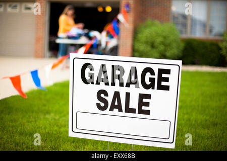 Garage Sale Schild an den Vorgarten ein Vorstadthaus mit einer Frau, die Elemente zu betrachten, auf einem Tisch. Stockfoto