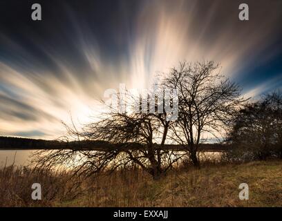 Schöne Langzeitbelichtung Landschaft des verschwommenen Himmel über verwelkte Apfelbaum am Seeufer. Schnell bewegte Wolken fotografiert auf lange Stockfoto