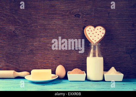 eine herzförmige Cookie und die Zutaten für Koch es, wie Milch, Eiern, Mehl, butter und Zucker auf einen blauen rustikalen hölzernen Vertikalfilter Stockfoto