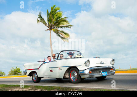 VARADERO, Kuba - 7. Juni 2011: Touristen fahren in weiße klassische amerikanische Oldtimer fahren auf eine Küstenstraße mit Palme. Stockfoto