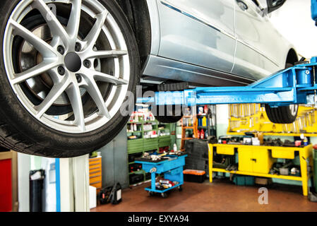 Auto auf einem Aufzug oder Hebezeug in einer Werkstatt oder Garage mit Fokus auf das Hinterrad und einen Blick auf die Werkzeuge in der Werkstatt Stockfoto