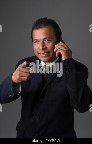 Hispanic Geschäftsmann hält eine Zelle Telefon, Blick in die Kamera und lächelnd zeigte mit einen Finger. Stockfoto