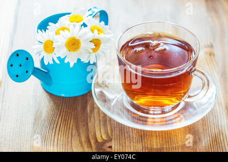 Kamillentee in eine Tasse und Kamille Glasblumen in eine kleine blaue Gießkanne Stockfoto