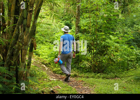 Einen jungen männlichen Erwachsenen ein Spaziergang durch den Regenwald in Bali, Indonesien. Tragen blaue Sport-Shirt, graue Hose, Hut, kam holding Stockfoto