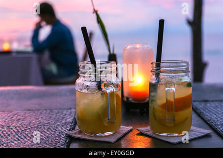 zwei mittelgroße Mojito-Gläser mit einem Strohhalm mit eine kleine gelbe Lampe hinter sich und eine Person im Hintergrund auf einen Tisch gestellt. Stockfoto