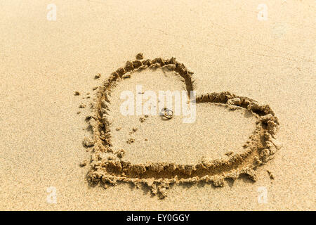 zwei goldene Hochzeit Tings auf einander platziert in der Mitte des Herzens, die am Strand in den Sand gezeichnet wird. horizontale b Stockfoto