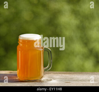 Pint goldene Bier auf rustikalen Holz mit unscharfen, grünen Bäumen im Hintergrund. Frisch gegossen, um die Natur zu genießen. Stockfoto