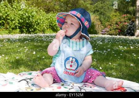 Kleines Mädchen mit einem Sonnenhut sitzt auf einem Teppich im Freien essen Brot Stockfoto