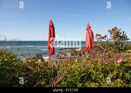 Das Fischerdorf Kikhavn. Blick auf das Kattegat Meer vorbei an Heckenrosen, Strand Terrassenmöbel und zwei rote Sonnenschirme an einem windigen Tag Stockfoto
