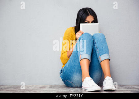 Lustige Frau, die auf dem Boden sitzend und bedeckte ihr Gesicht mit Tablet-PC auf graue abckground Stockfoto