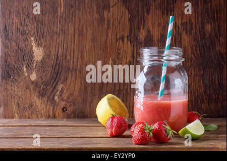 Glas mit roten Erdbeer Smoothie, serviert mit frischen Erdbeeren, Zitrone und Limette auf Holztisch Stockfoto