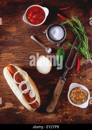Hausgemachte Hot Dog mit Senf Zutaten, Tomatensauce, Zwiebel, Chili-Pfeffer, Rosmarin. Auf Holztisch mit Fleischgabel und Löffel Stockfoto