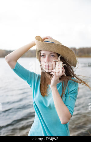 Sommersprossige Mädchen mit Hut eine Shell anhören und stehend auf dem See bei windigem Wetter Stockfoto