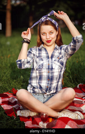 Junge schöne lächelnde Frau, gekleidet in Pin-Up Stil sitzt auf einer roten Plaid auf dem grünen Rasen im Sommerpark Stockfoto