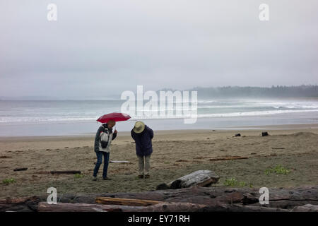 Älteres Paar zu Fuß am Strand von Wickanninish an einem regnerischen Sommertag.  Surfer in den Wellen des Ozeans in der Ferne. Long Beach, BC Stockfoto