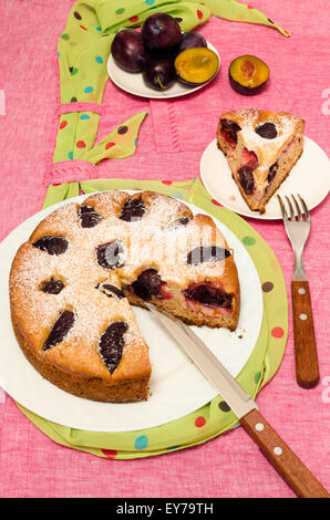 Hausgemachte Kuchen mit Pflaumen auf ein rosa Tischdecke. Stillleben mit Besteck, rustikalen Stil. In der Nähe von Messer, Gabel und frisches Obst. Stockfoto