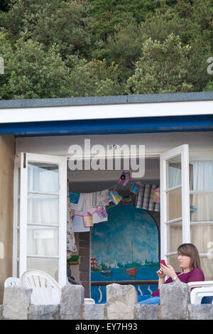 Junge Frau mit Telefon sitzen draußen bunte Strandhütte auf Promenade zwischen Canford Cliffs und Sandbänken, Poole im Juli Stockfoto