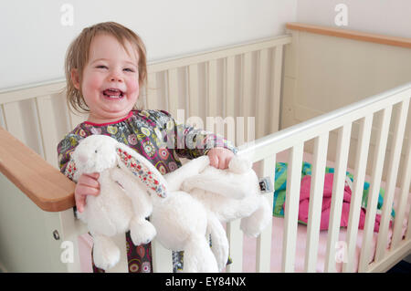 Glückliche kleine Mädchen stehen in ihrem Kinderbett mit Plüschtieren spielen Stockfoto