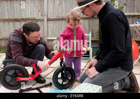 Kleine Mädchen beobachten ihr Großvater und Vater ihr rotes Laufrad montieren Stockfoto