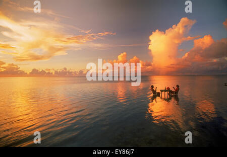 Pärchen, die am Tisch sitzen und einen Drink bei Sonnenuntergang in der Lagune inmitten des Pazifischen Ozeans genießen. Cook-Inseln. Urlaub Ruhe und Einsamkeit im Paradies Stockfoto