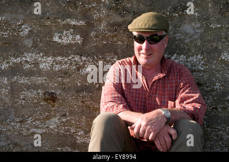 Porträt von einem mittleren Alter Mann mit Sonnenbrille und eine Mütze, sitzen gegen eine Steinmauer Stockfoto