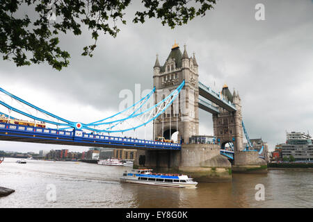 Tower Bridge, London, umrahmt von Bäumen an einem grauen Tag und ein Touristenboot auf dem Fluss. Stockfoto
