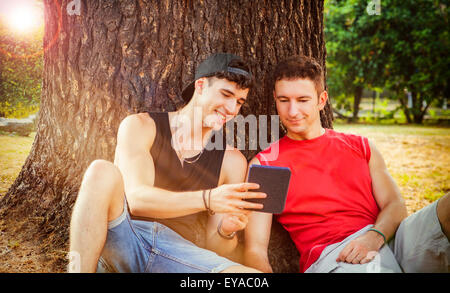 Zwei junge männliche Freunde mit lässig trendige Sommerkleidung stützte sich auf einen alten Baumstamm, während gerade etwas lustiges auf einem Tisch Stockfoto