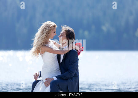 Der Bräutigam hat seine Braut auf den Knien, am Ufer eines Sees. Sie Lächeln romantisch zueinander. Stockfoto