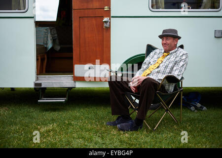 Mann, mit Hut und Krawatte, sitzt auf einem Campingstuhl neben seinem Oldtimer-Wohnwagen auf einer Autolampe in Ripon, North Yorkshire. VEREINIGTES KÖNIGREICH Stockfoto
