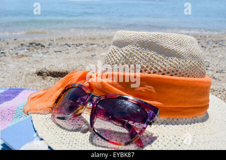 Strohhut-Sonnenbrille und ein Buch mit Meer im Hintergrund an einem sonnigen Tag am Strand Stockfoto