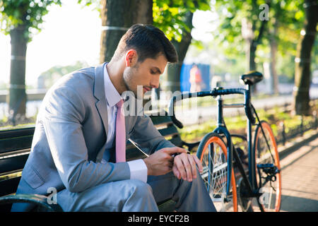 Junger Geschäftsmann sitzt auf der Bank im Freien und Blick auf die Armbanduhr Stockfoto