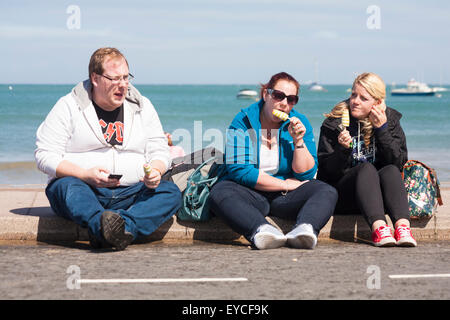 Drei Leute sitzen auf Bürgersteig essen Eis am Stiehl in Swanage im Juli Stockfoto