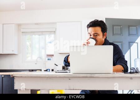 Reife Geschäftsmann Tippen auf Laptop Whist trinken Kaffee zum Mitnehmen am Küchentisch Stockfoto