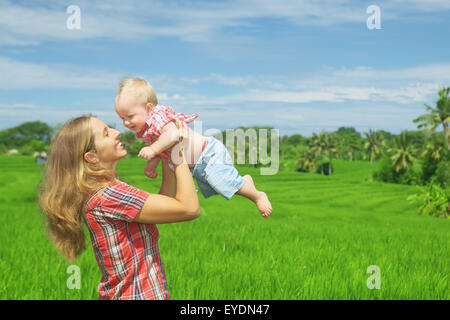 Glückliche Mutter warf sich fröhliche Baby Sohn auf balinesischen grüner Reis Terrassen Hintergrund. Im freien gesundes Kind Lebensstil Stockfoto