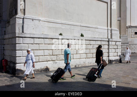 Zwei Nonnen gehen vorbei an Touristen mit Gepäck in der Nachmittagshitze unter den Mauern der Kirche Santa Maria della Salute in Dorsoduro, di Stockfoto