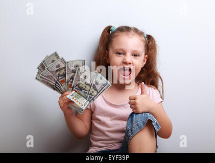 Glücklich genießen Kind Mädchen halten Geld und zeigt Daumen oben Zeichen auf blauem Hintergrund Stockfoto