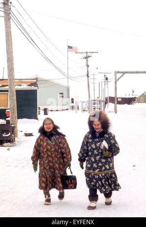 Das Tragen schwerer Wintermäntel, gehen zwei Frauen in Führungspositionen Eskimo auf einer Straße in Barrow, Alaska, an einem verschneiten Wintertag. Beachten Sie die amerikanische Flagge am Postamt im Hintergrund. Stockfoto