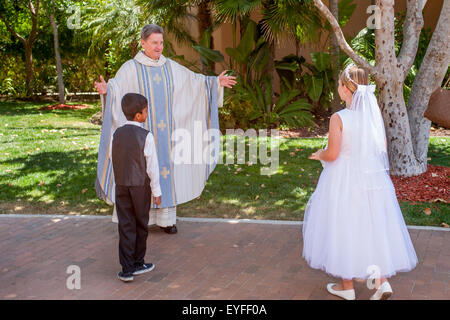 In formalen Outfits, multirassische Kinder Roben Monsignore einer Laguna Niguel, CA, katholischen Kirche nach Erstkommunion Zeremonien begrüßen. Erste Heilige Kommunion ist auch bekannt als das Sakrament der Heiligen Eucharistie. Stockfoto