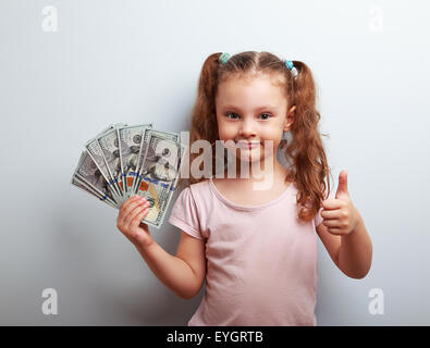 Glücklich Rich kid Girl halten Geld und zeigt Daumen oben Zeichen auf blauem Hintergrund mit leeren Textfreiraum Stockfoto