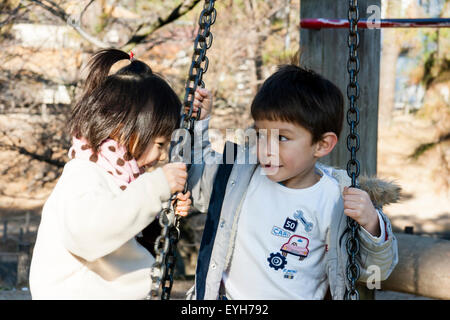 Zwei Kinder, Junge und Mädchen, sitzend auf Schaukeln, suchen sehr Coy, während Sie chatten und an einander. Junge Kaukasier, Mädchen ist Japanisch. 4-5 Jahr. Stockfoto