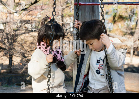 Zwei Kinder, Junge und Mädchen, sitzend auf Schaukeln, suchen sehr Coy, während Sie chatten und an einander. Junge Kaukasier, Mädchen ist Japanisch. 4-5 Jahr. Stockfoto