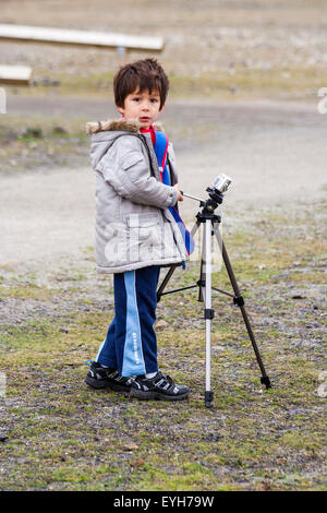 Junge mit einer Kamera auf einem Stativ im Freien auf grobe Anschluss montiert. Kaukasier Kind, 6-7 Jahre, braune Haare, trägt Mantel und Schal. Winter, Landschaft. Stockfoto