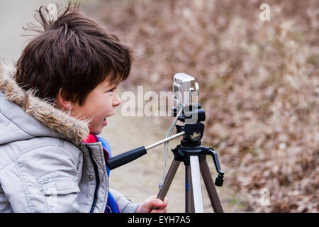 Junge mit einer Kamera auf einem Stativ im Freien auf grobe Anschluss montiert. Kaukasier Kind, 6-7 Jahre, braune Haare, trägt Mantel und Schal. Winter, Landschaft. Stockfoto