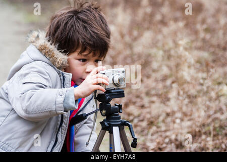 Junge mit einer Kamera auf einem Stativ im Freien auf grobe Anschluss montiert. Kaukasier Kind, 6-7 Jahre, braune Haare, trägt Mantel und Schal. Winter, Landschaft. Stockfoto