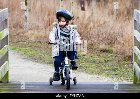 Junge Reiten Fahrrad mit Stabilisatoren, auf groben Track im Winter. Kaukasier Kind, 6-7 Jahre alt, mit Mantel und Helm. Holzzäune auf beiden Seiten. Stockfoto